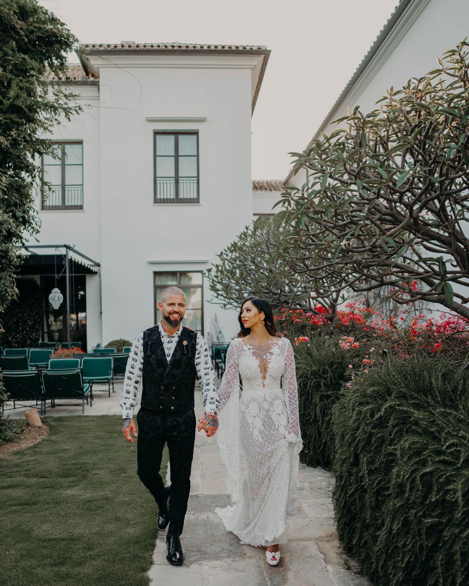 Elegant newlyweds walking along a white Mediterranean villa with blooming gardens and blue outdoor seating.
