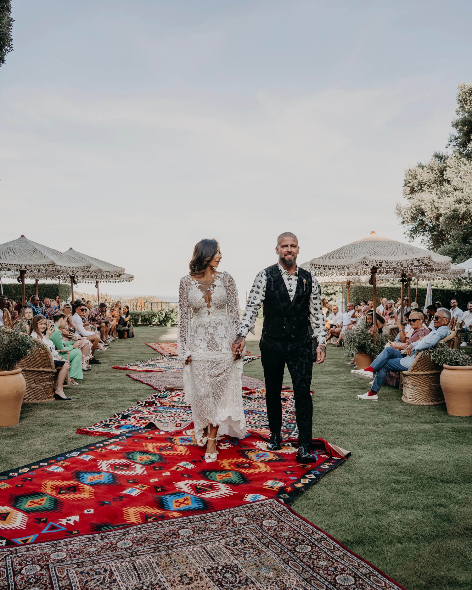 A couple walks on their wedding aisle decorated with a vibrant red Persian rug surrounded by guests on a lawn.