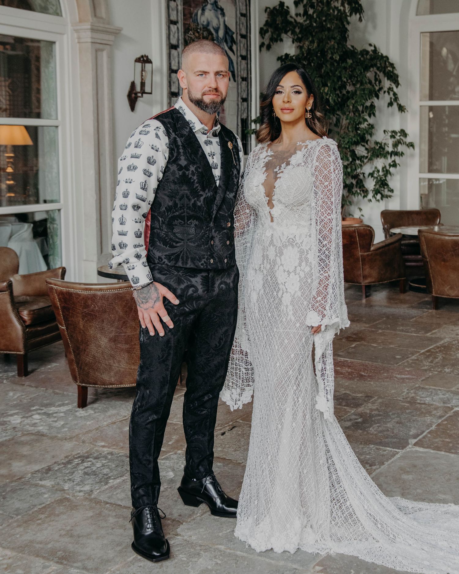 Elegant couple poses in formal wedding attire featuring a lace gown and black patterned vest.