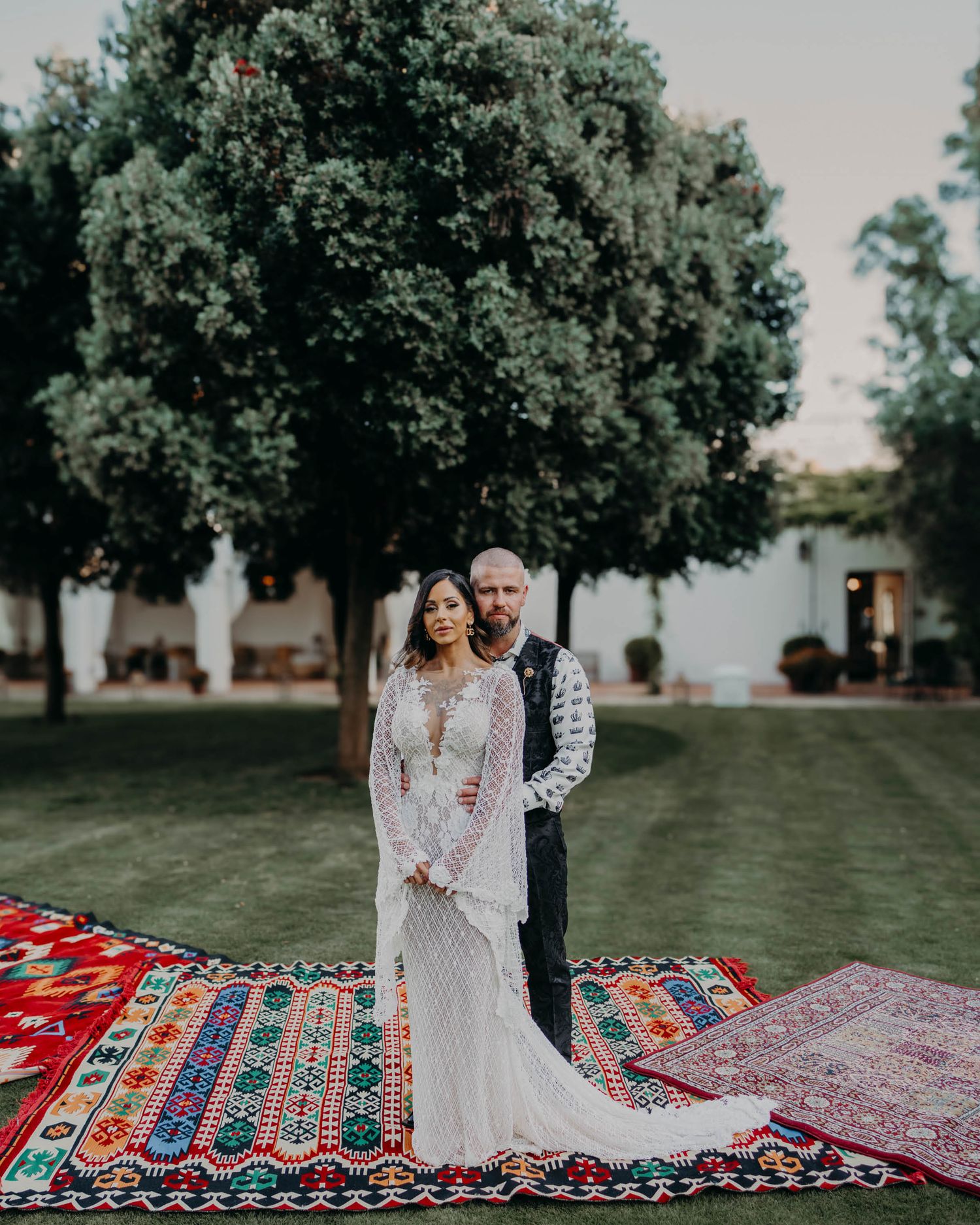 Couple stands together on vibrant layered Moroccan rugs under large green trees during outdoor wedding ceremony.