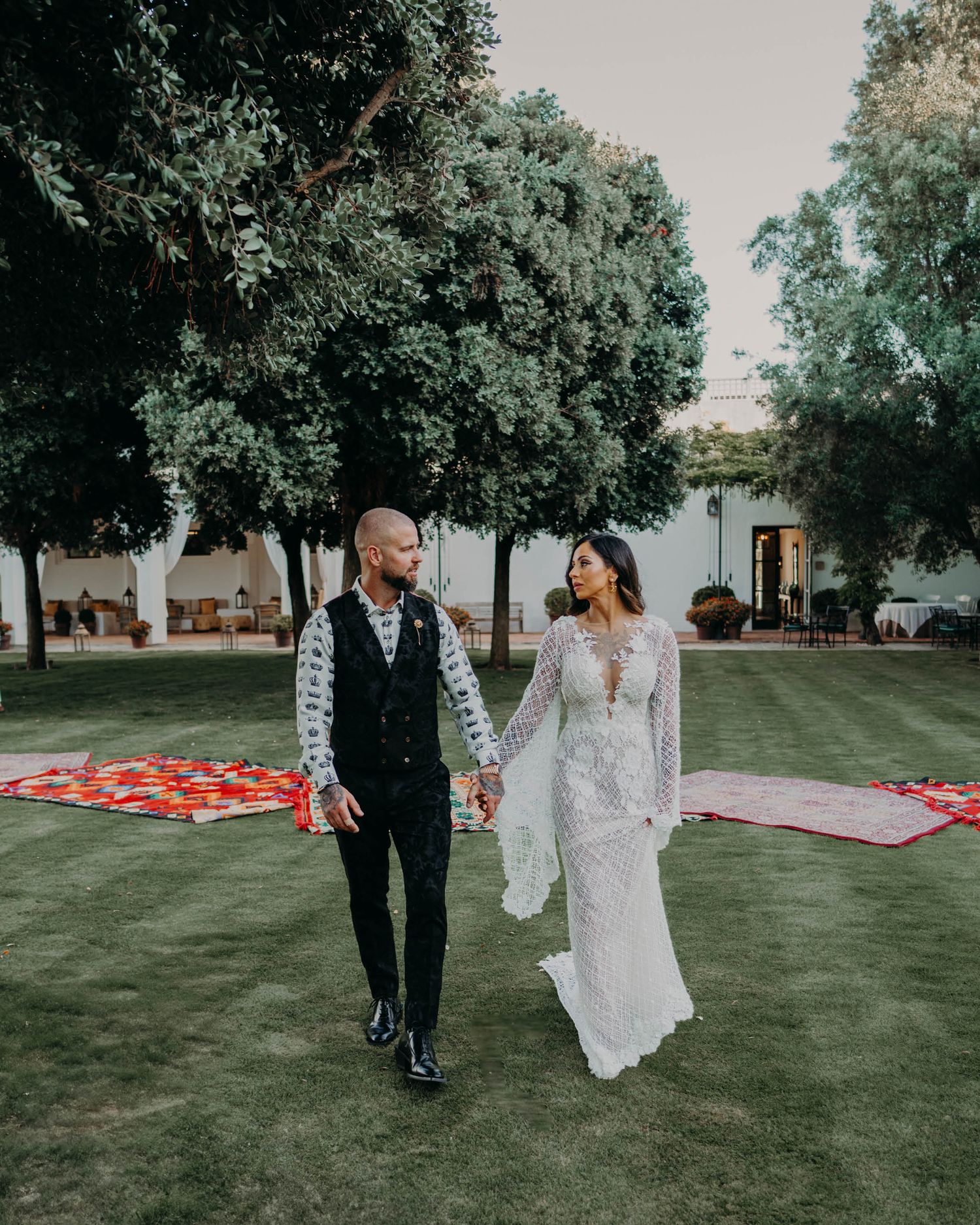 A romantic wedding couple walks hand in hand on a lawn with decorative rugs and lush trees in the background.