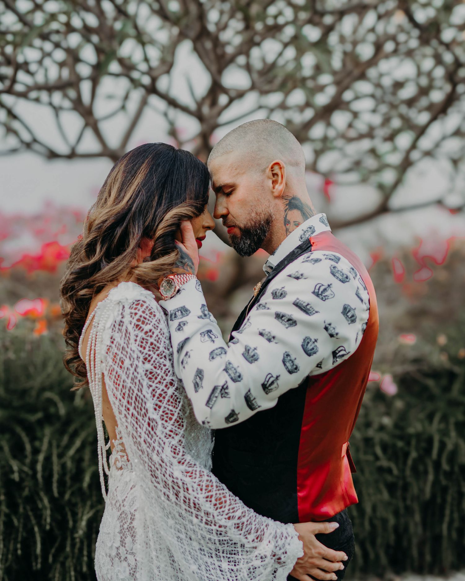 A romantic outdoor portrait shows a couple in a sweet embrace among blurred red flowers and twisting tree branches.
