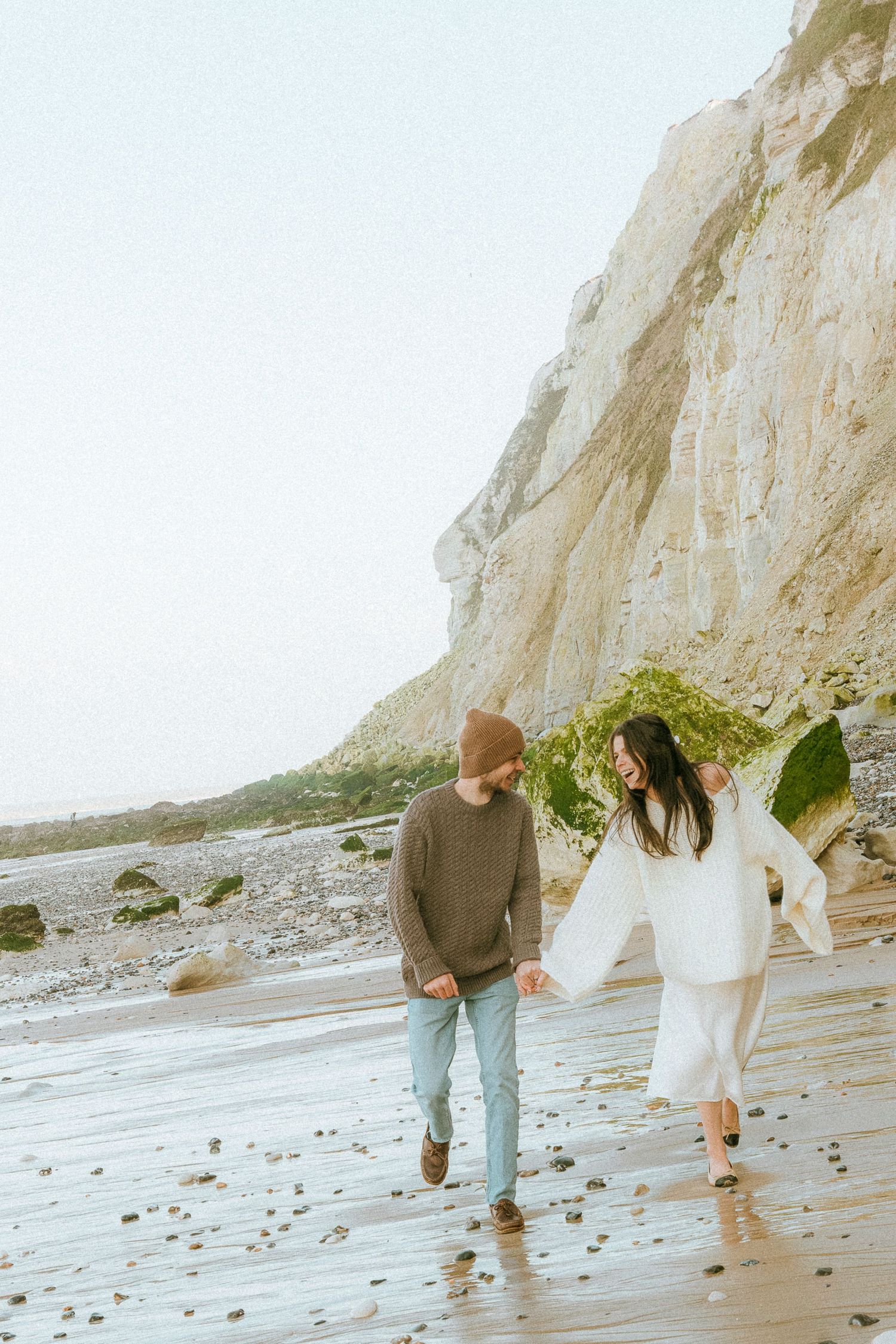 A couple walks barefoot along a misty beach beneath towering white cliffs on an overcast day.