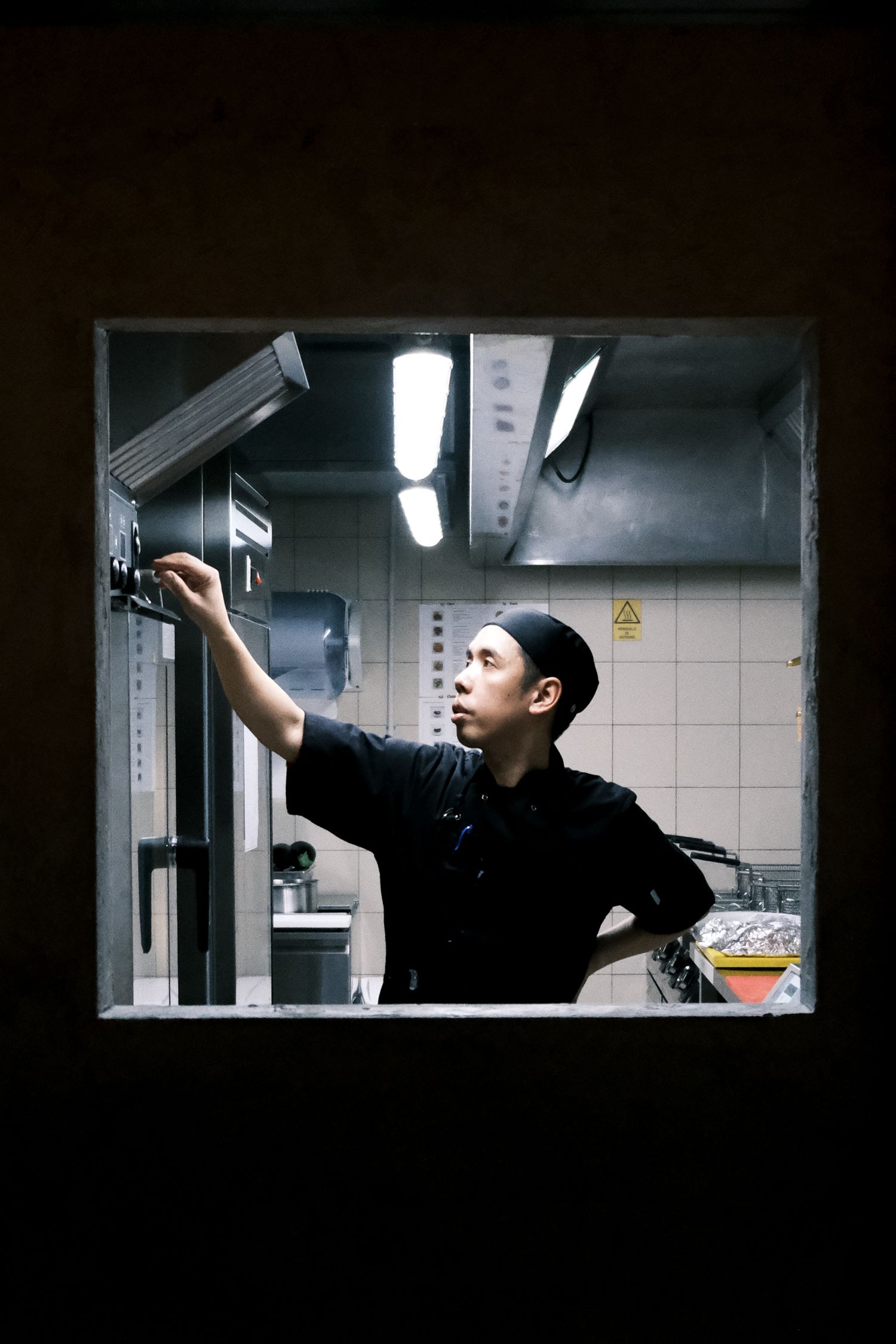 Kitchen worker reaching up to adjust equipment through service window in dimly lit commercial kitchen.
