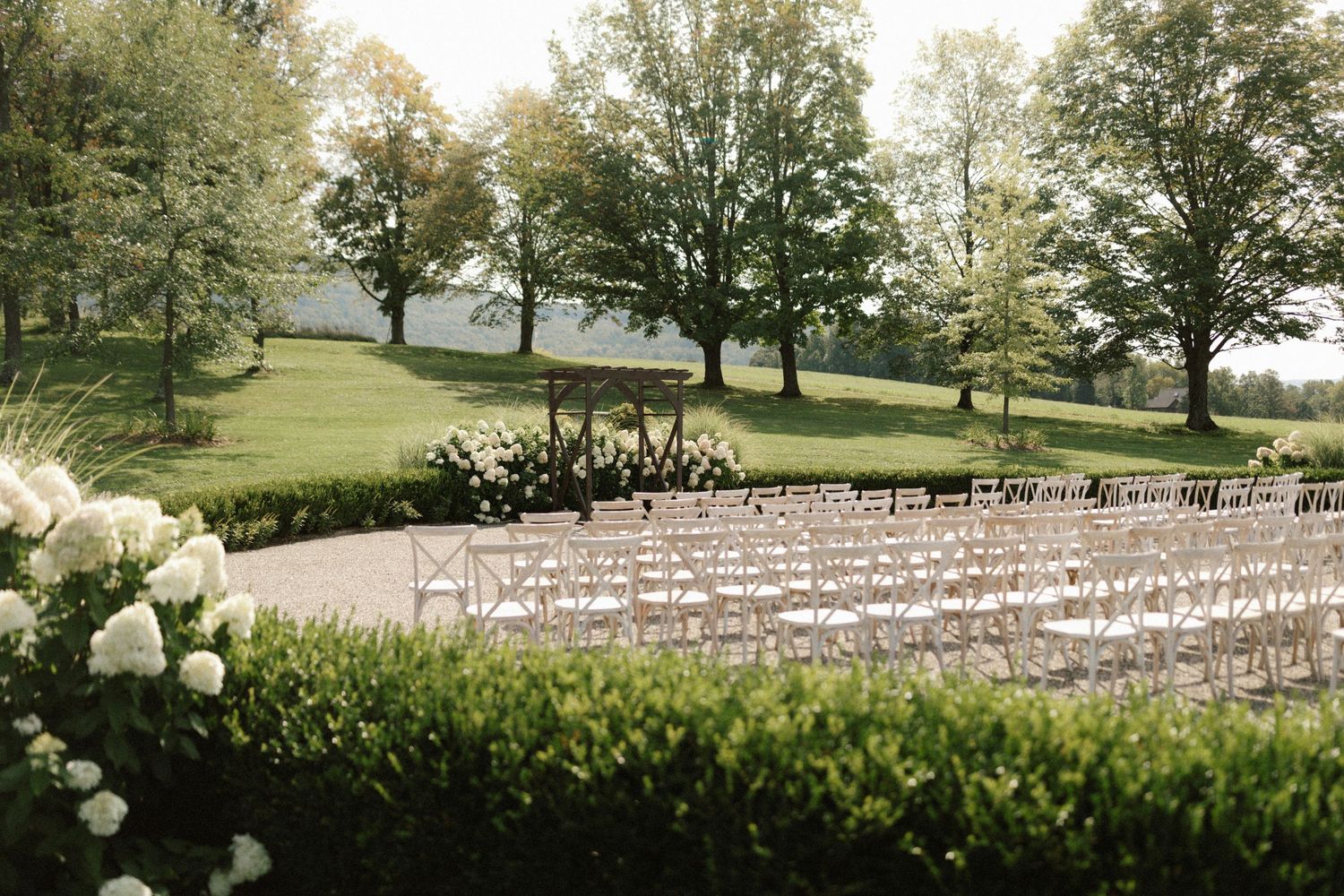 A scenic outdoor wedding ceremony setup with white chairs arranged on a lush lawn surrounded by trees and white floral decor.