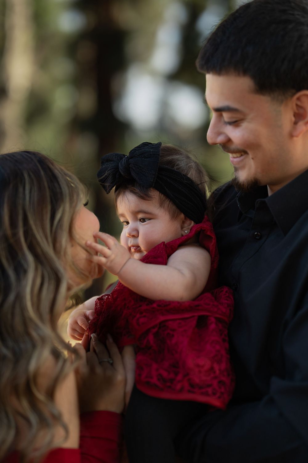 A baby in a red velvet dress shares a tender moment with family during an outdoor photo session.