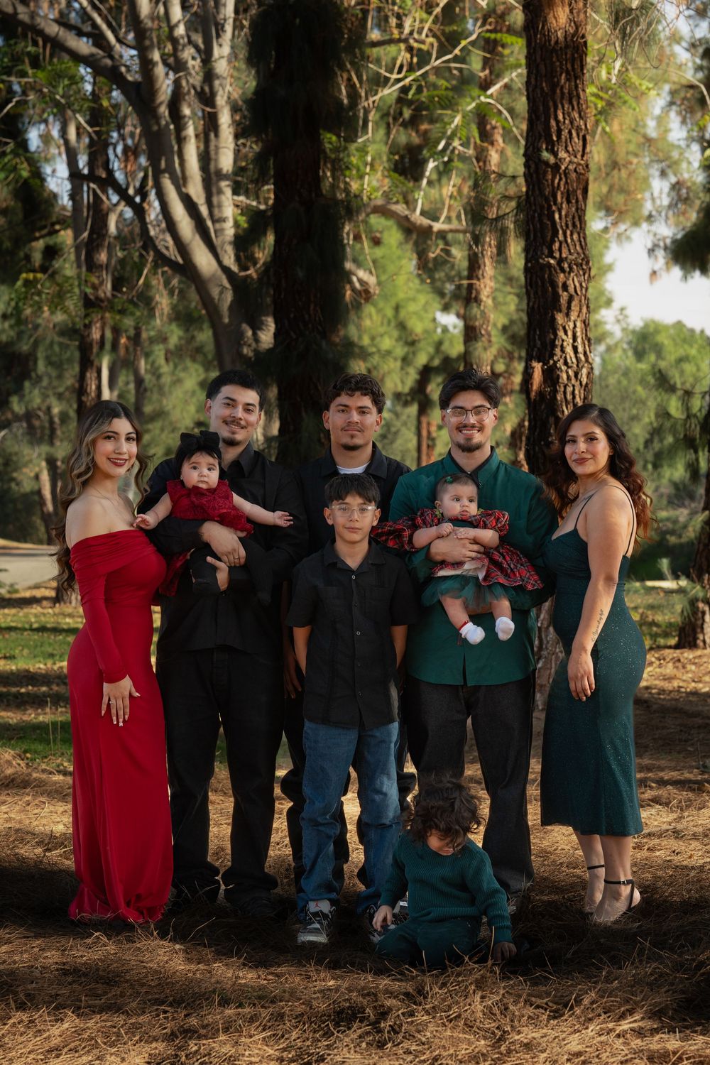 A group poses together outdoors against large tree trunks in coordinated green and red outfits during a family photoshoot.