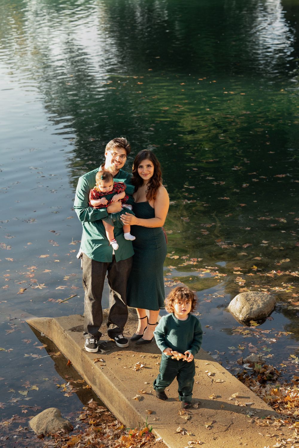 A family stands together on a stone ledge by a pond with autumn leaves floating on the water's surface.