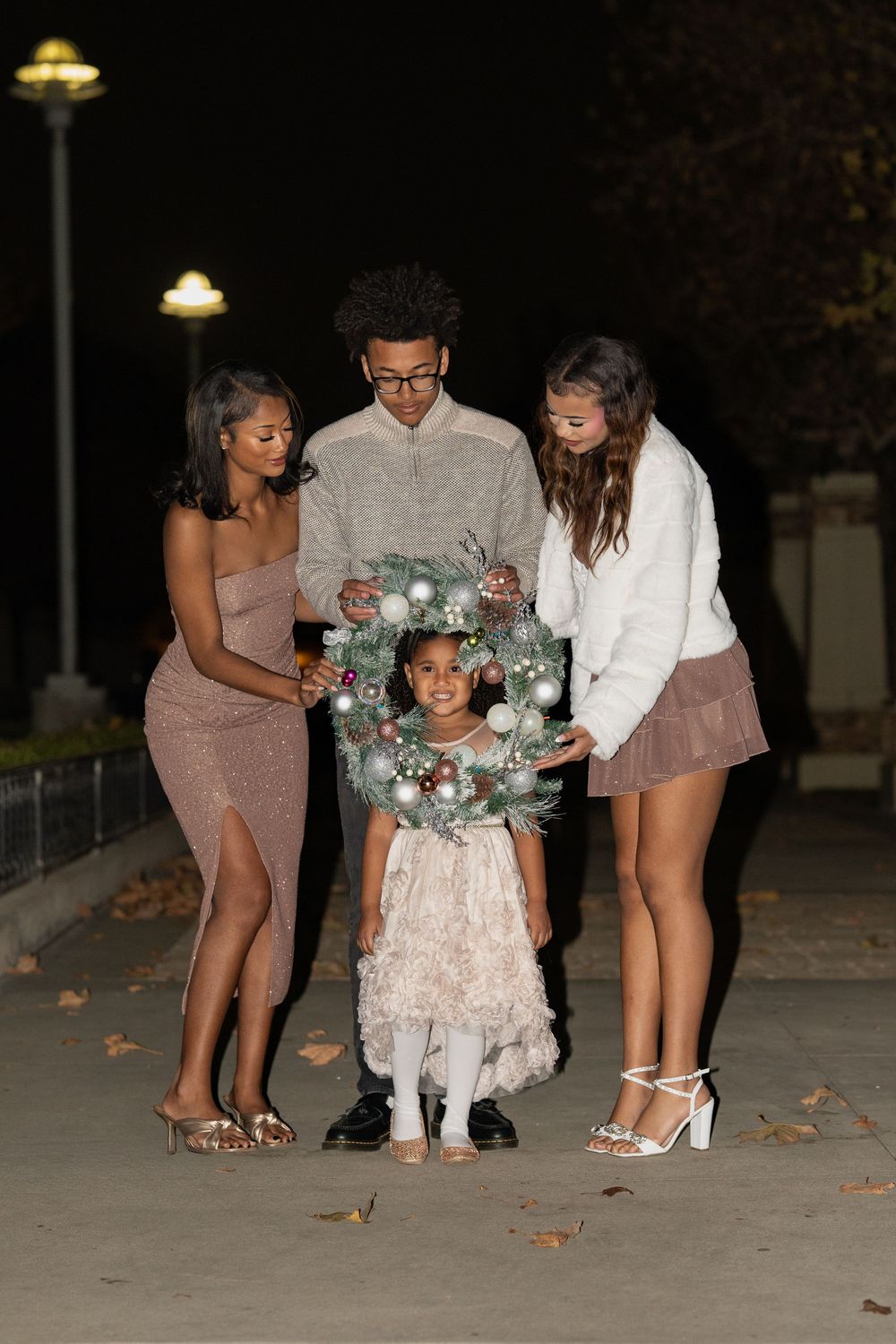 A group of friends gather around a young child holding a festive holiday wreath at night under lamp posts.