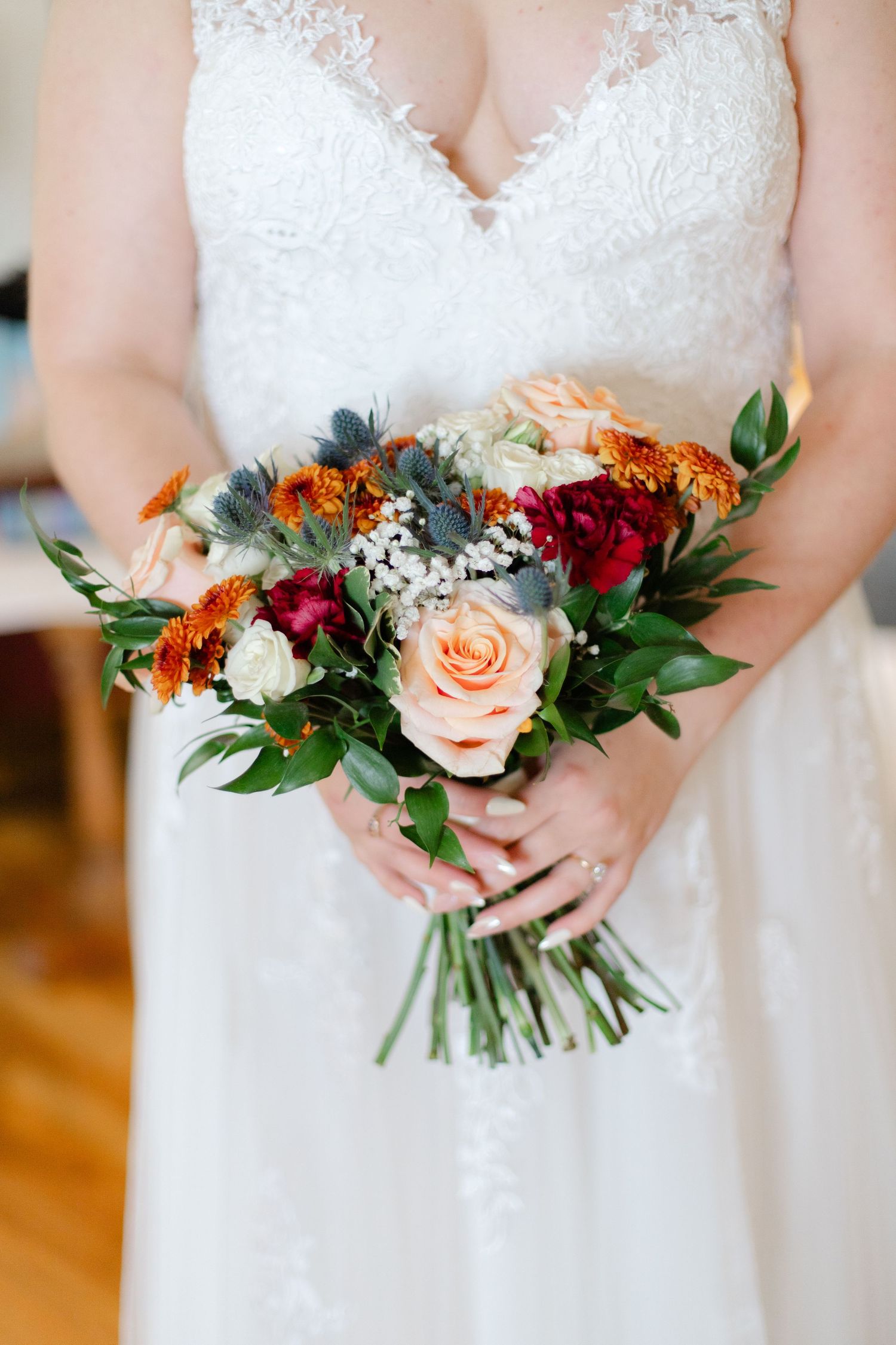 Bride in a lace white wedding dress holds colorful bouquet with peach roses, red flowers, and greenery.
