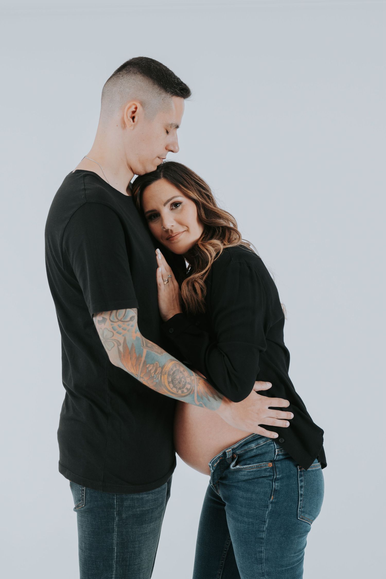 Couple in casual black outfits embracing during intimate photoshoot against grey backdrop.