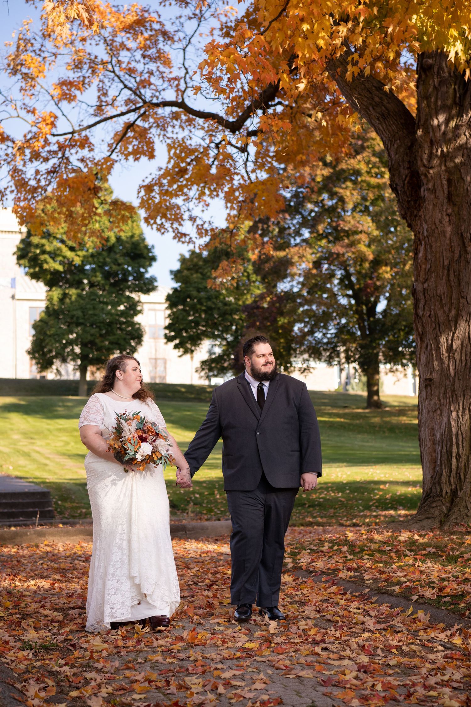 A couple in wedding attire stands on a path surrounded by fall foliage with brilliant orange autumn leaves overhead.