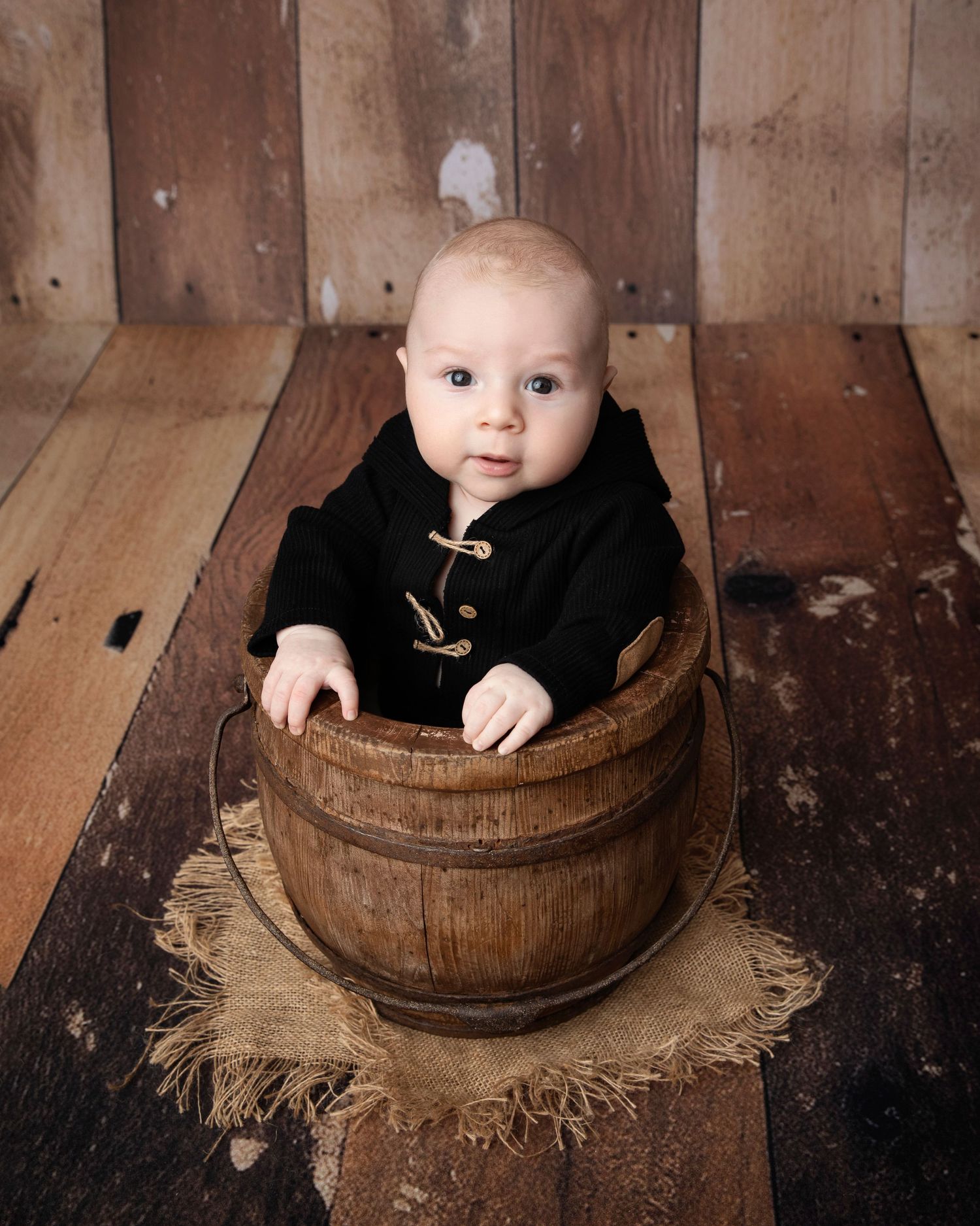 Small child in black outfit sitting inside rustic wooden barrel against wooden wall background.