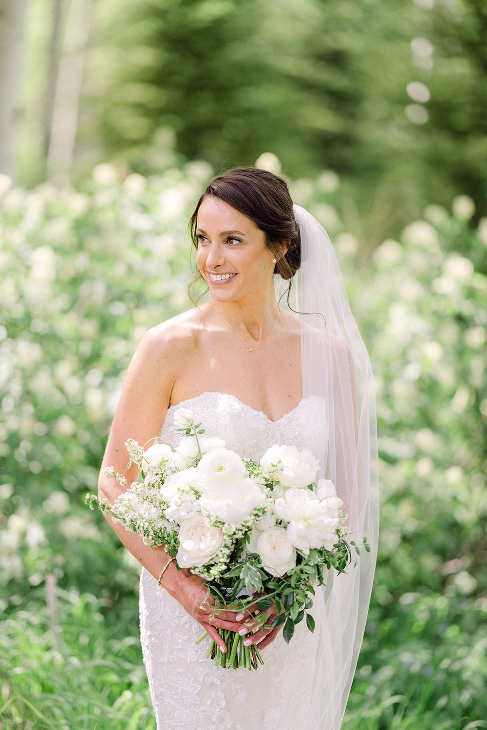 Bride in strapless lace gown holds white peony bouquet amidst mountain wildflowers.