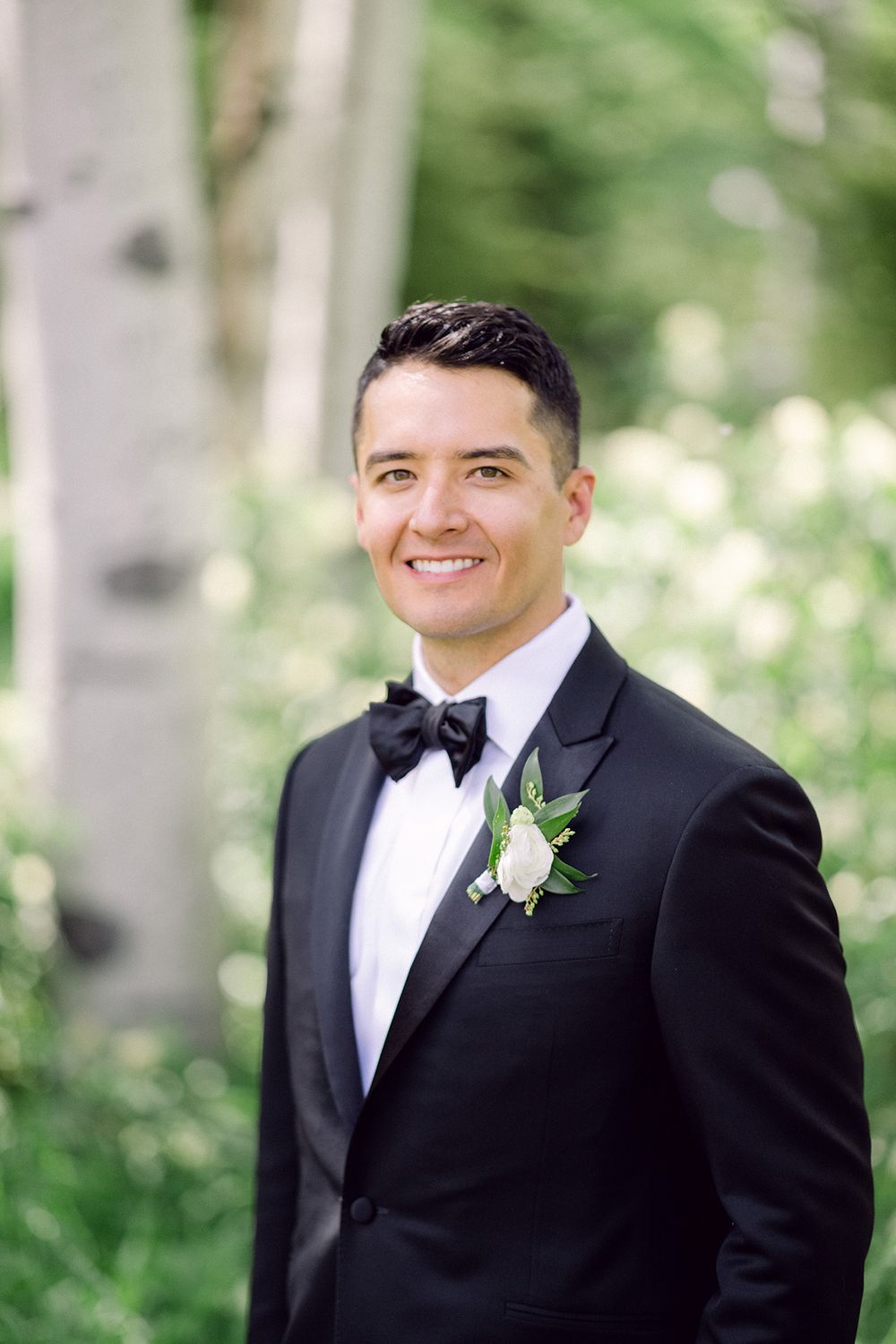 Groom in classic black tuxedo with white boutonniere smiles in mountain setting.