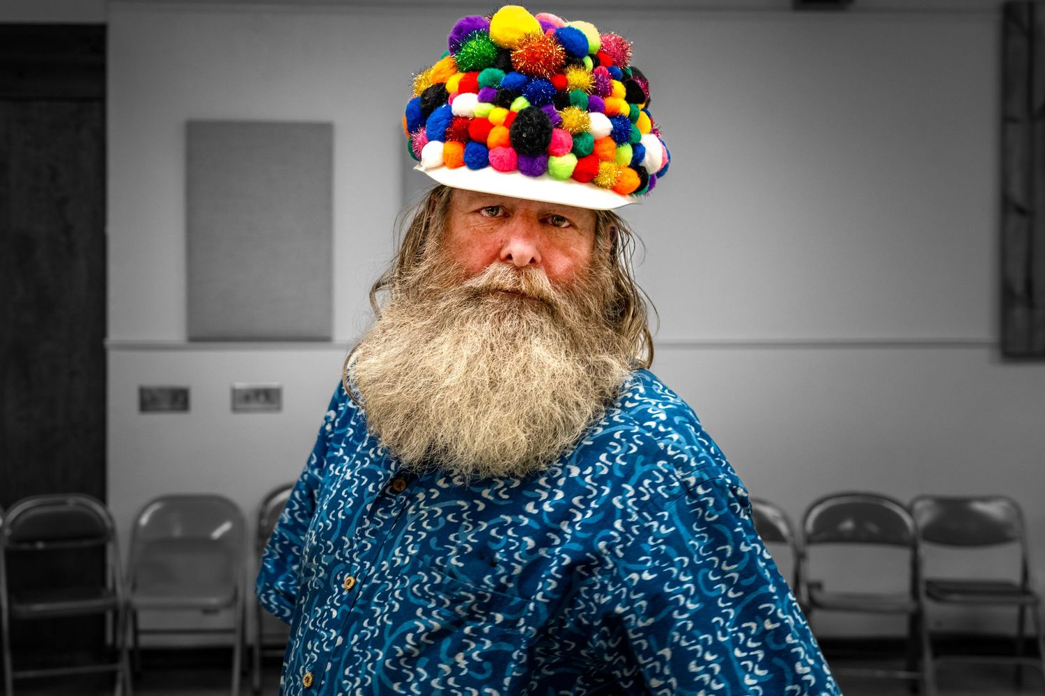 Person wearing colorful pom-pom hat and blue knit sweater against grey background.