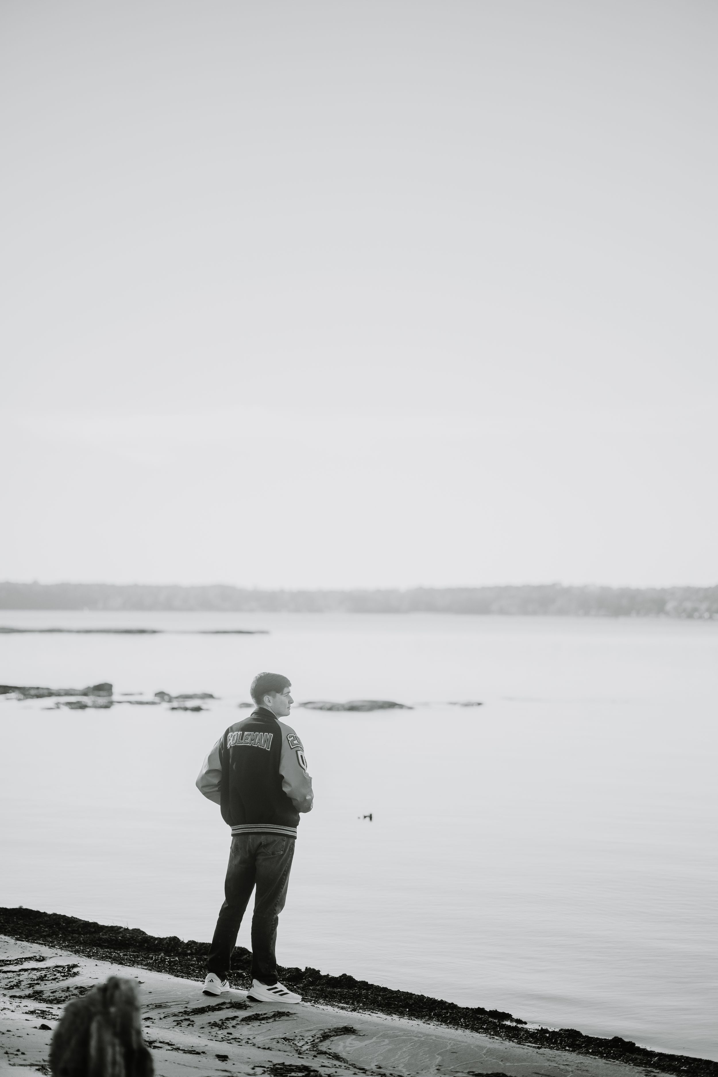 A black and white photograph of a person standing on a rocky shoreline overlooking a misty lake.