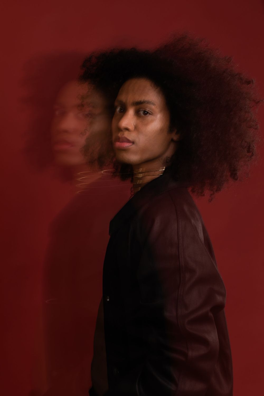 Artistic portrait against red background featuring flowing natural curly hair and burgundy leather jacket.