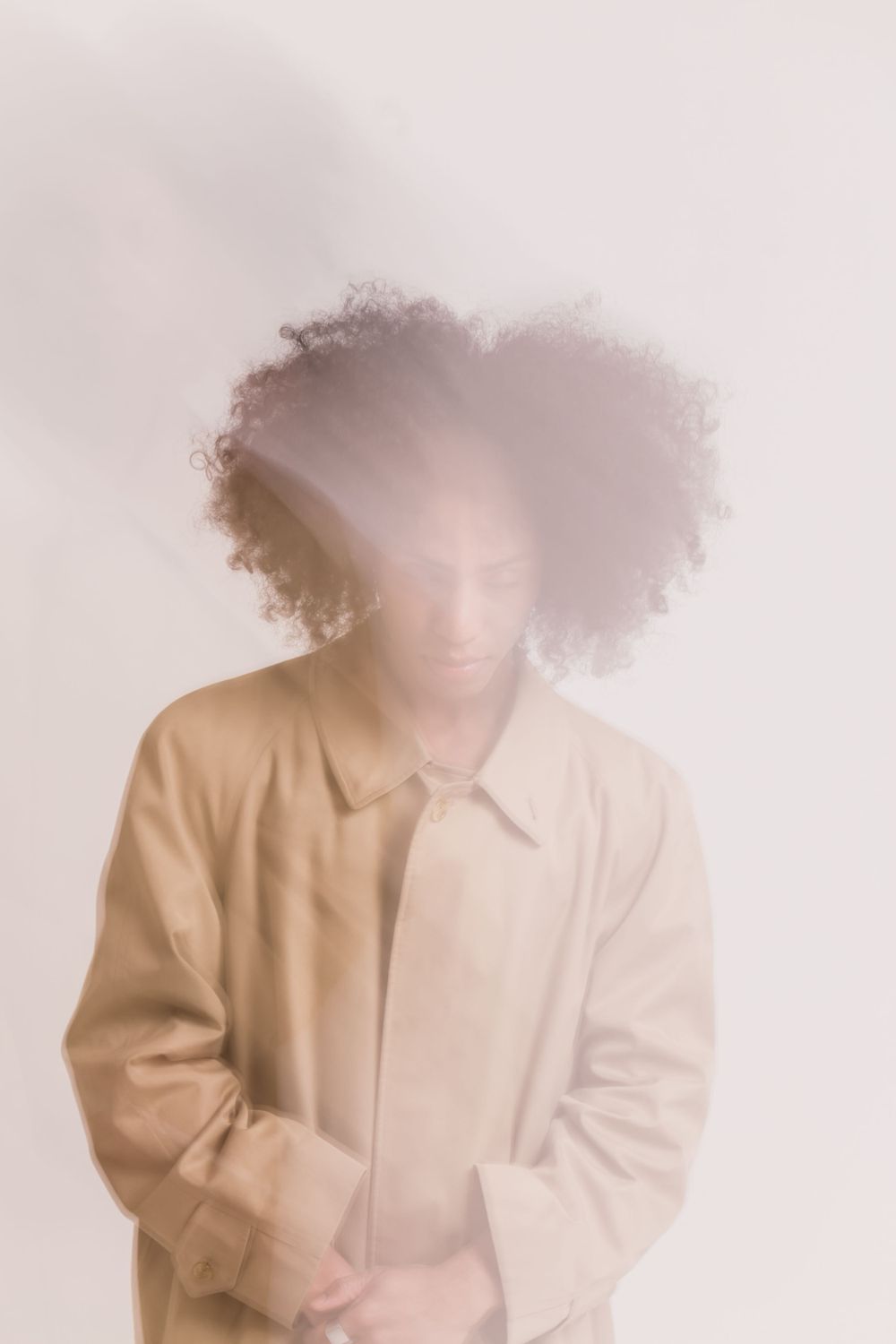 Ethereal portrait in soft lighting wearing a beige coat with natural hair against white background.