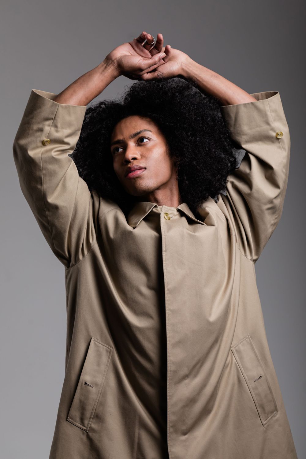 Fashion portrait with natural curly afro hair wearing beige oversized trench coat against gray studio backdrop.