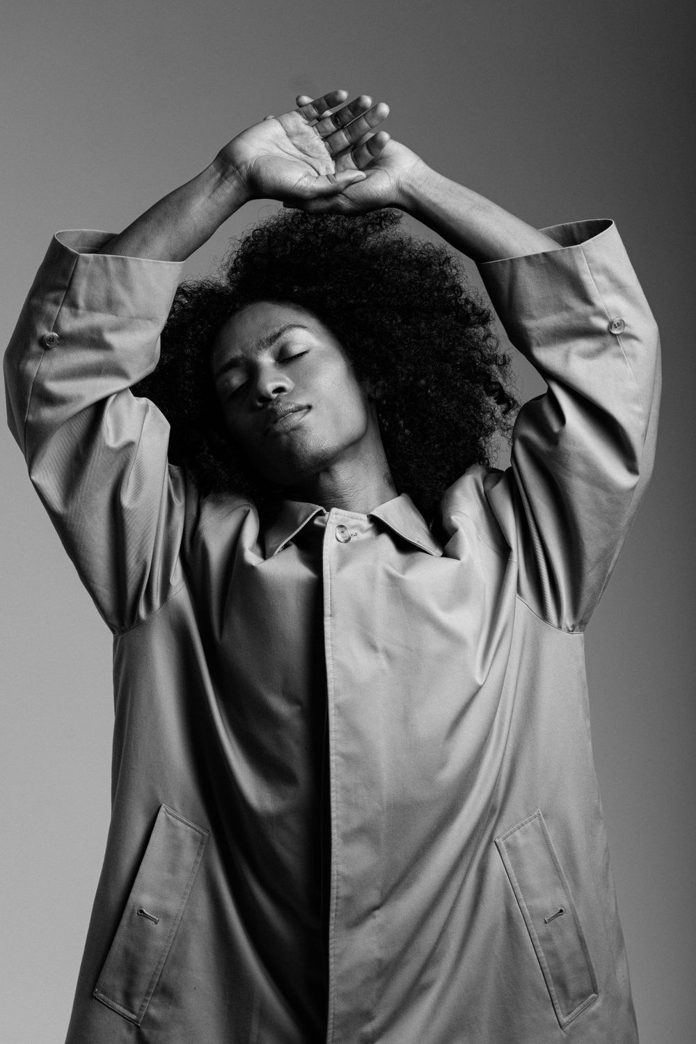 Artistic black and white portrait of person with natural curly afro hair stretching arms up while wearing light coat.