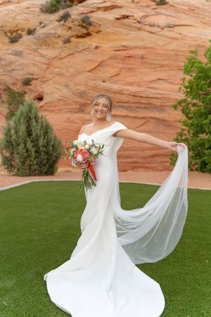 A flowing white wedding dress twirls gracefully on green grass against dramatic red rock formations.