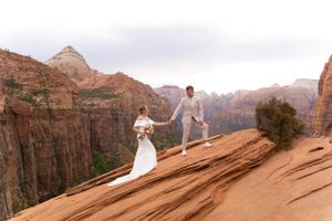 A wedding couple poses on red rock formations overlooking a scenic desert canyon landscape.