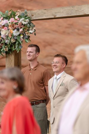 Wedding ceremony guests and participants gather under floral-decorated wooden posts against red rock cliffs.
