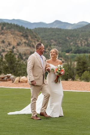 Wedding couple walks across a green lawn with mountain views in the background.