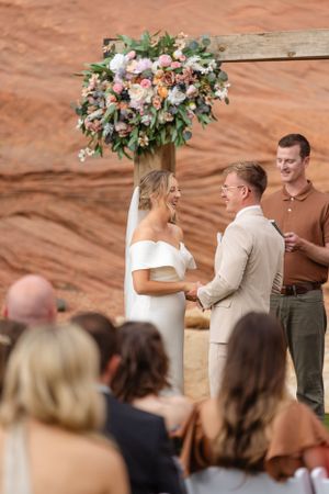 Wedding ceremony taking place under wooden posts with floral decorations against red rock backdrop.