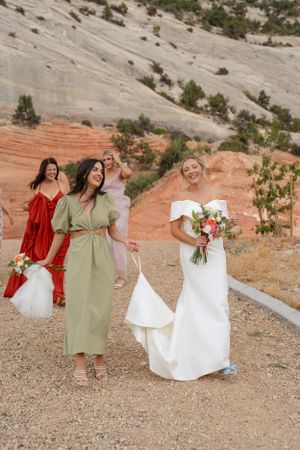 Wedding party members in green and white dresses laugh together against a scenic red rock backdrop.