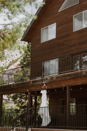 White wedding dress hanging on rustic wooden cabin balcony with forest backdrop.