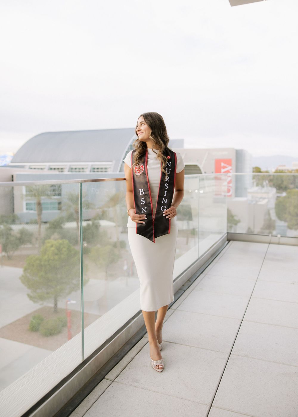 Person in white dress and patterned vest stands on modern balcony overlooking cityscape.