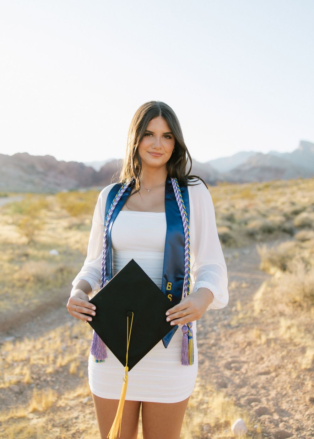 Graduate in white dress holds cap with graduation stole against desert backdrop.