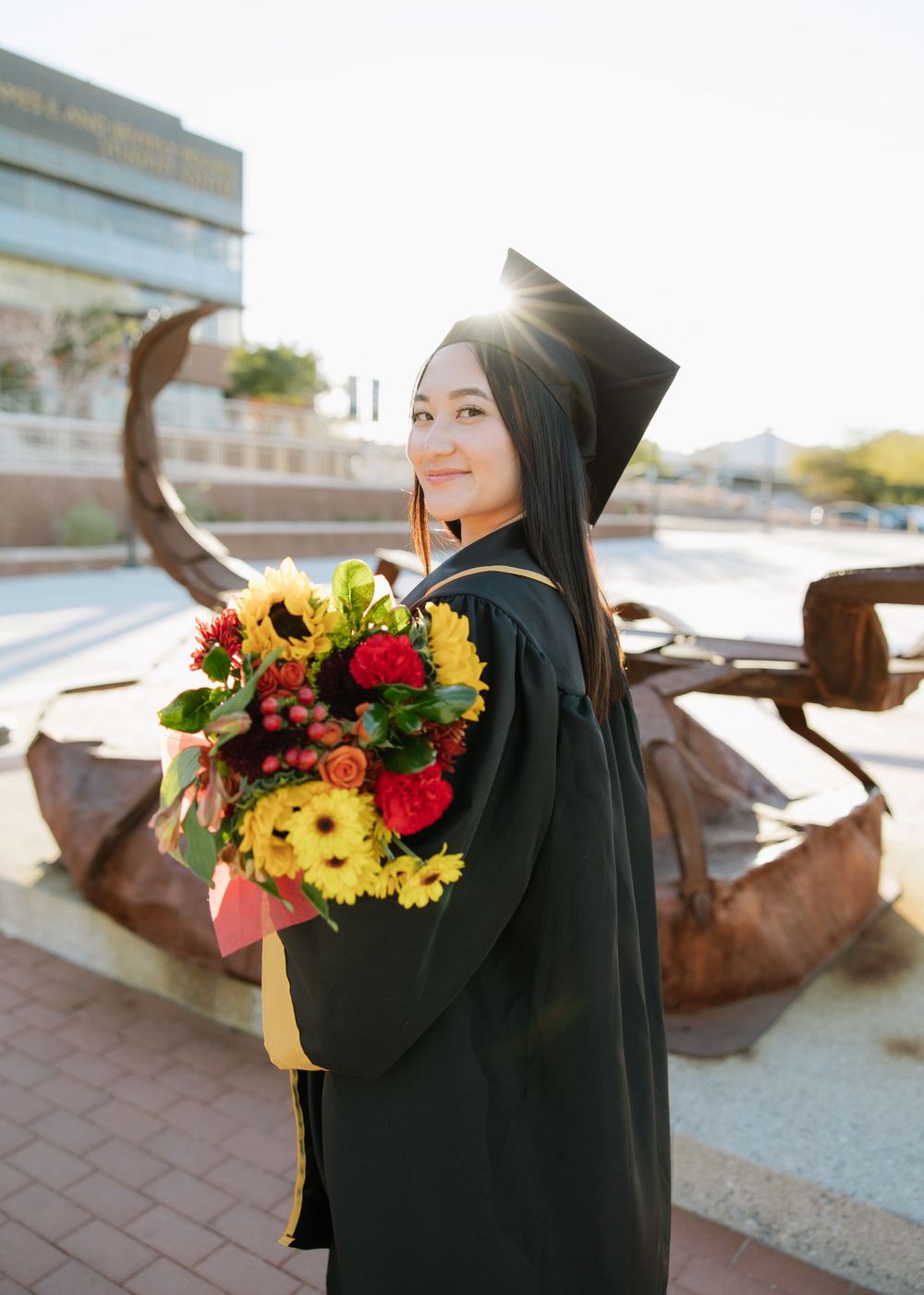 Graduate holding vibrant bouquet of sunflowers and roses in front of campus sculpture.