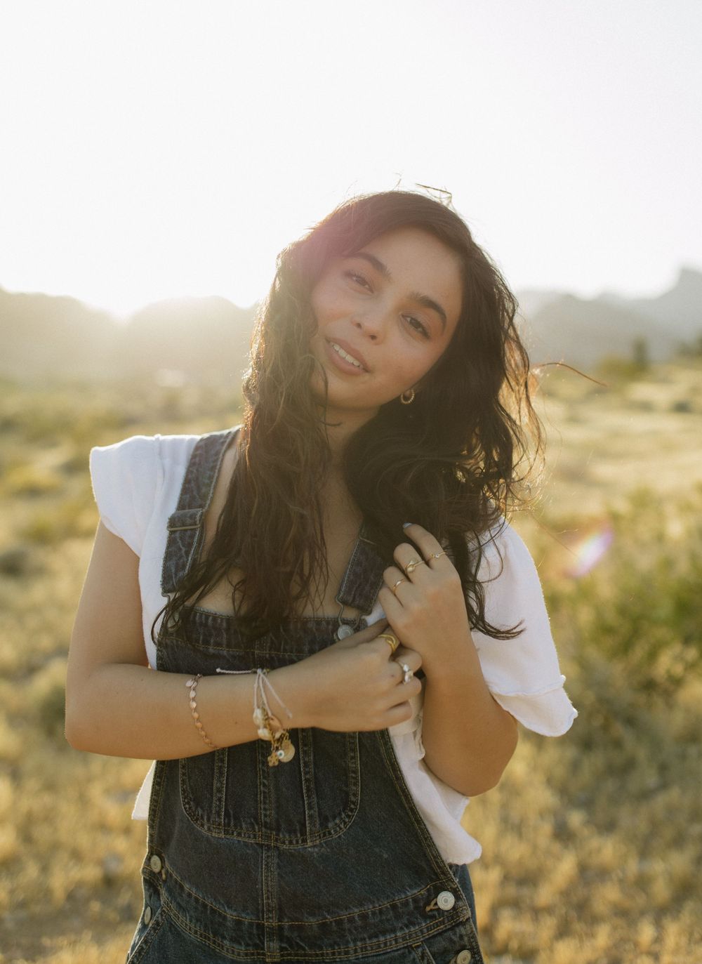 Person in denim overalls and white shirt poses during sunset in desert landscape.