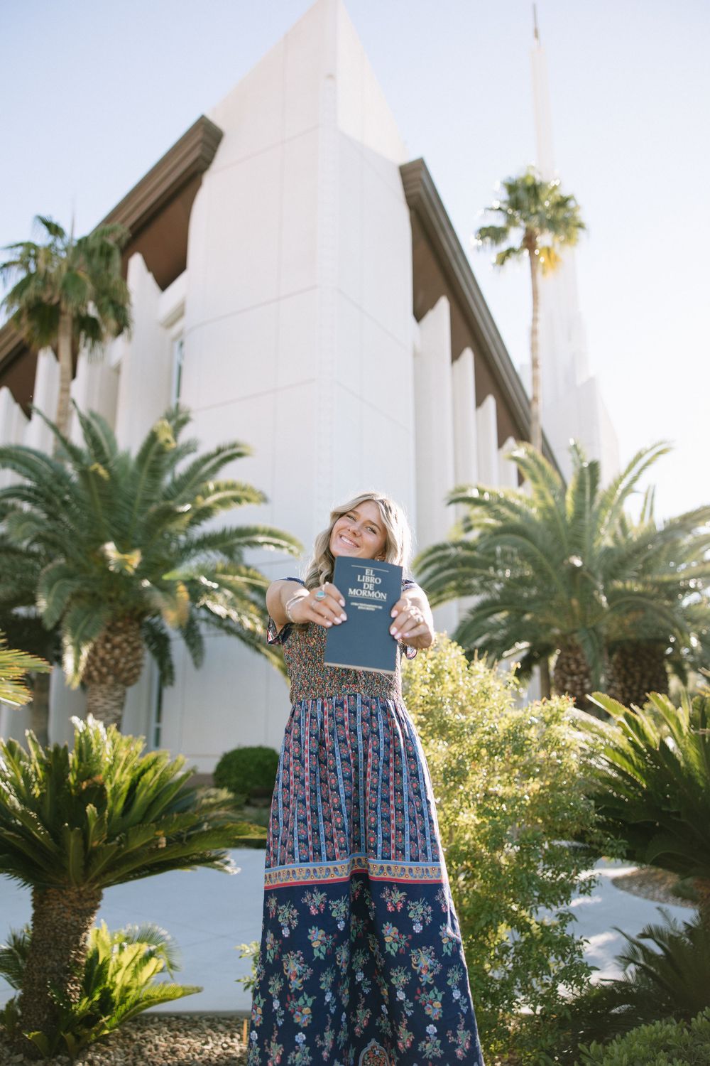 Person in colorful maxi dress poses in front of white church building with palm trees.