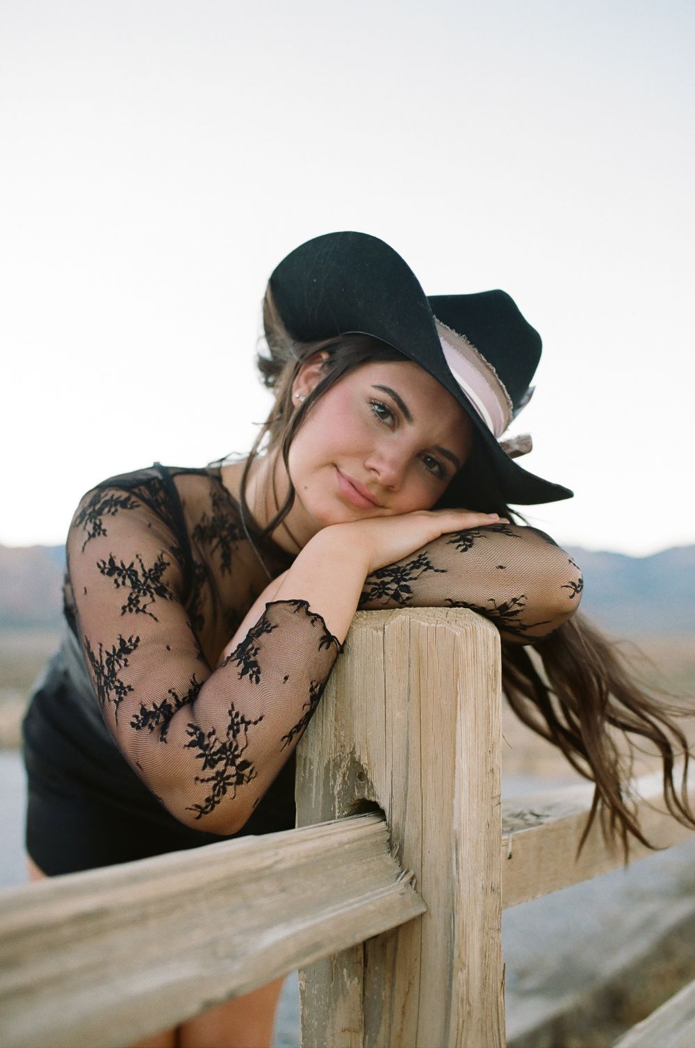 Woman in black hat and lace dress leaning on rustic wooden fence in desert landscape at sunset.