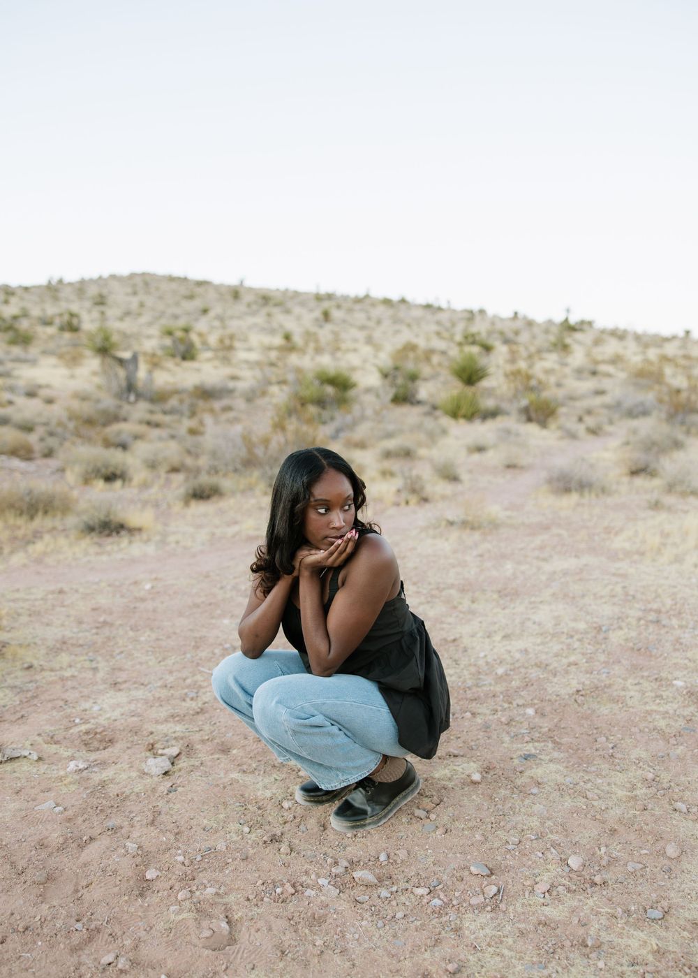 Person in casual attire crouching in desert terrain during a photo shoot.