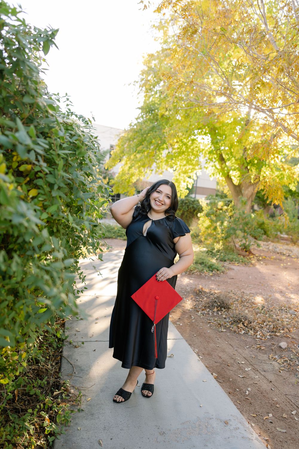 Graduate in black dress holding diploma and smiling on walkway surrounded by autumn trees and greenery.