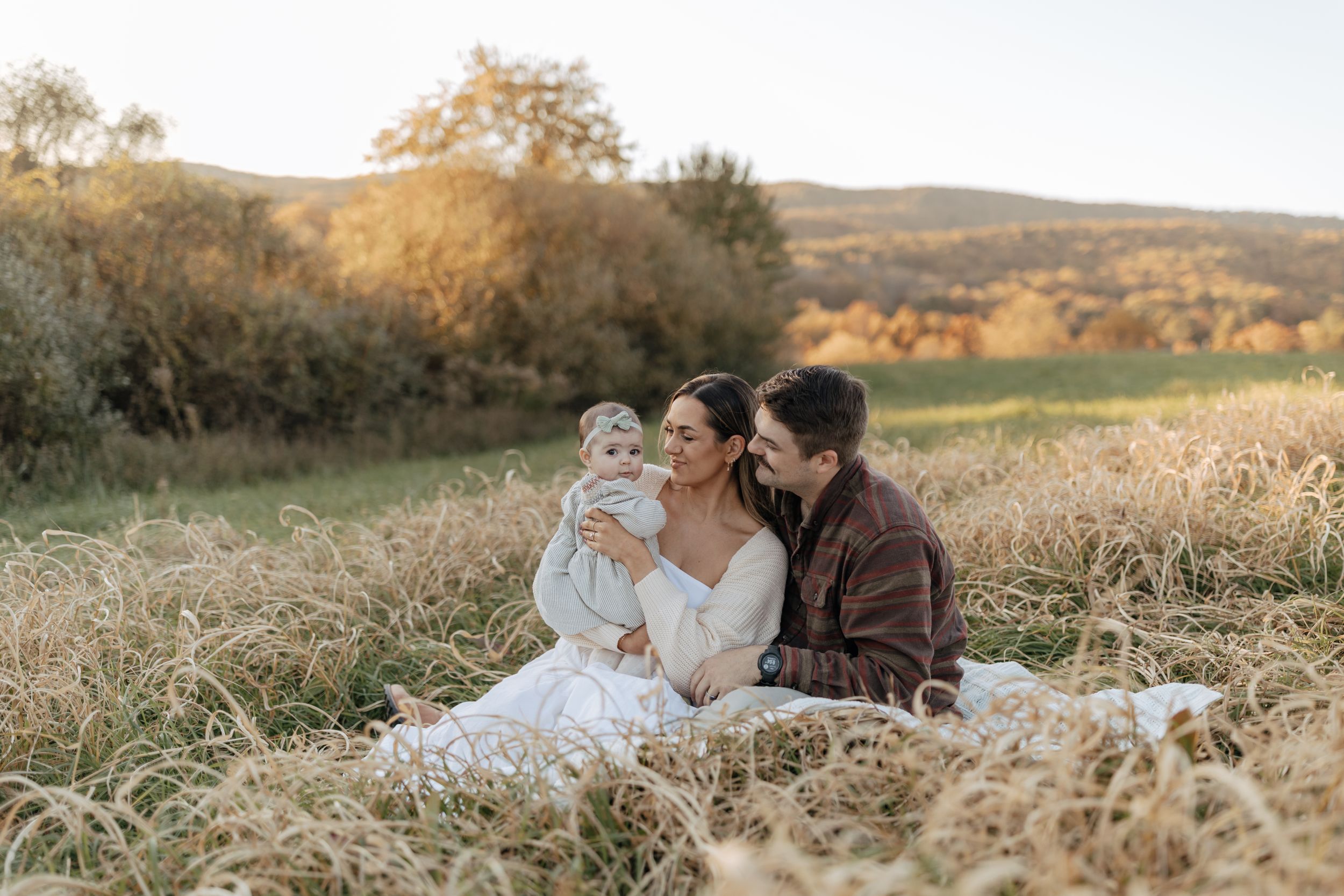 A sweet family moment captured in tall grass with autumn foliage in the background.