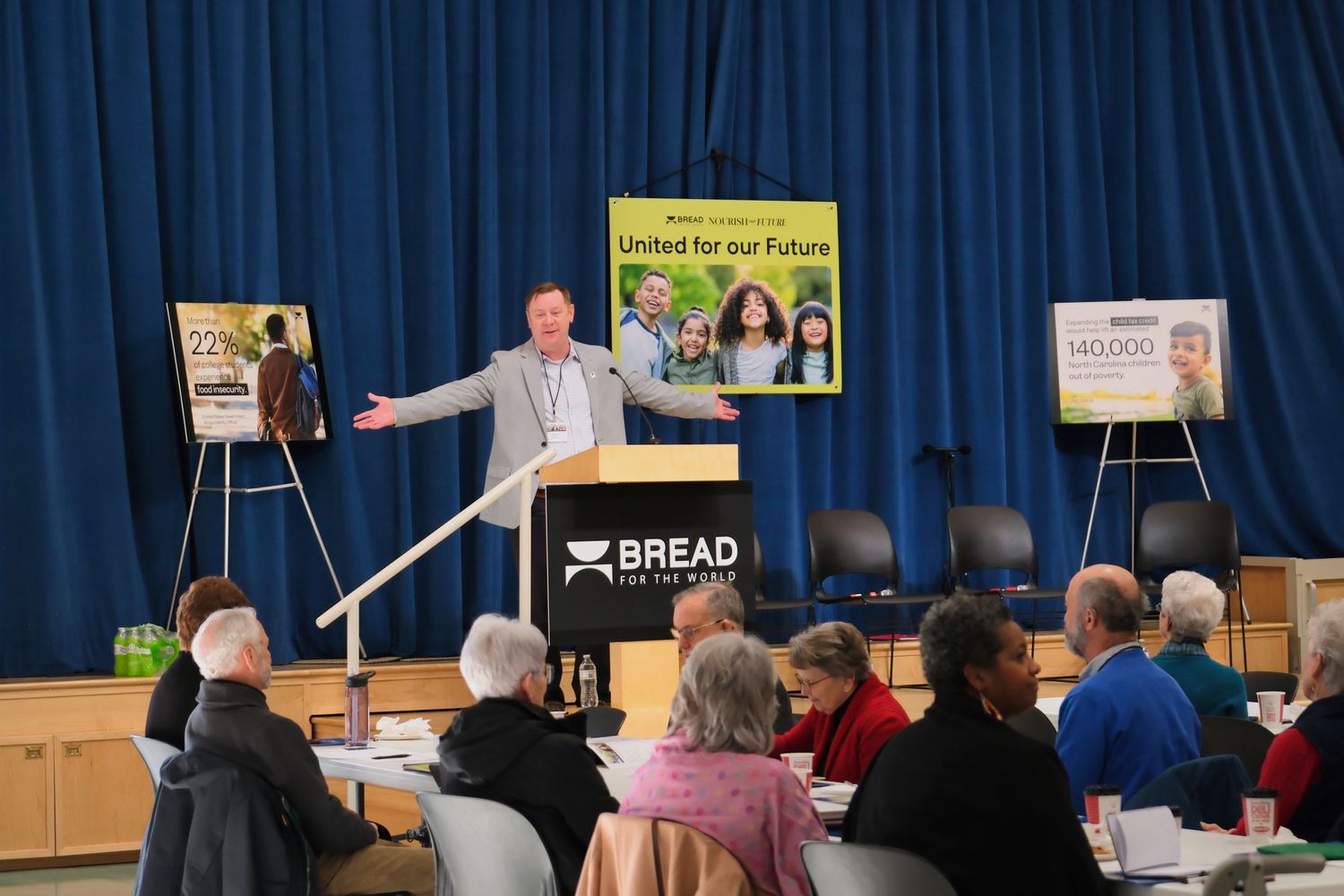 A presenter speaks to an audience in front of display boards at a Bread for Life program in a community center.