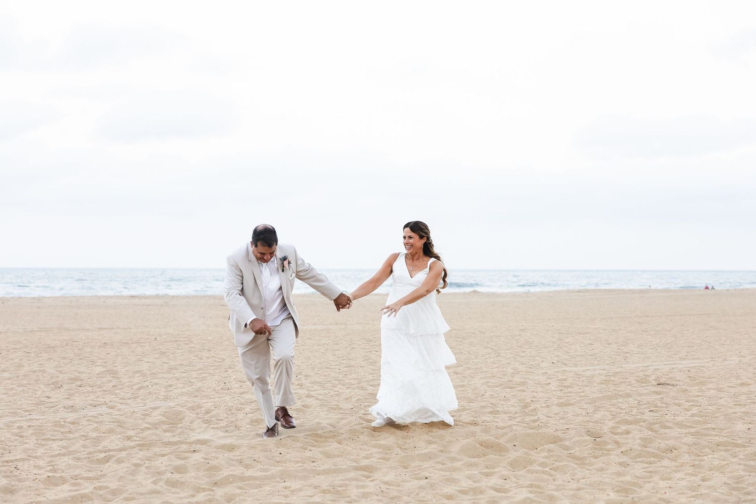 A couple in wedding attire holds hands while walking on a serene beach with cloudy skies.