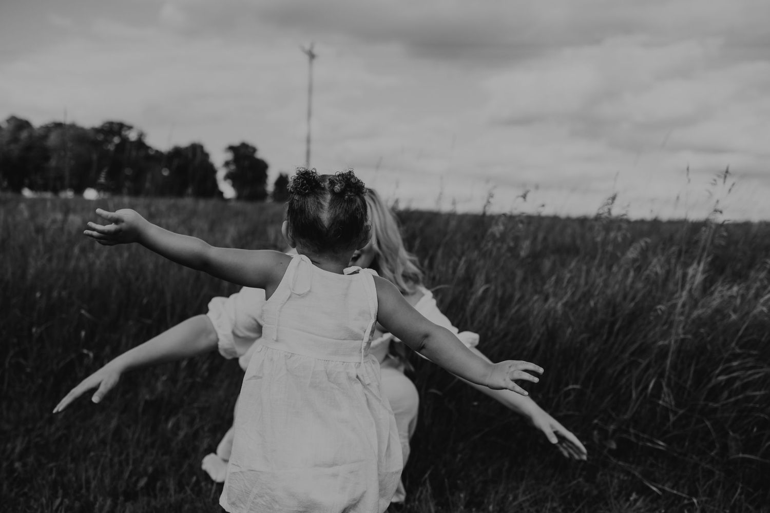 A figure in a white dress spins joyfully in a meadow of tall grass against an overcast sky in black and white.