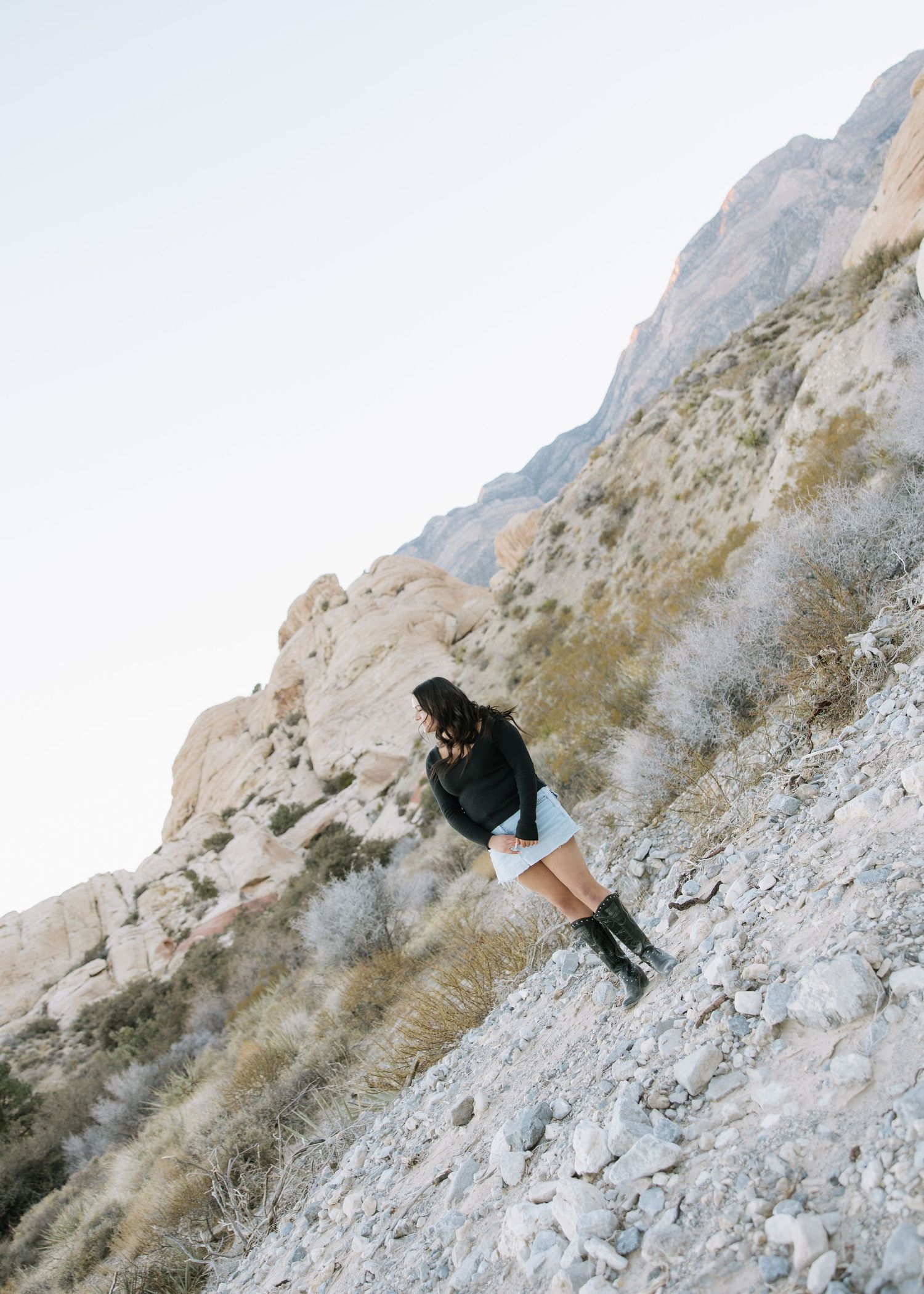 A person in a black sweater and white shorts climbs up a rugged mountainside trail in warm sunlight.