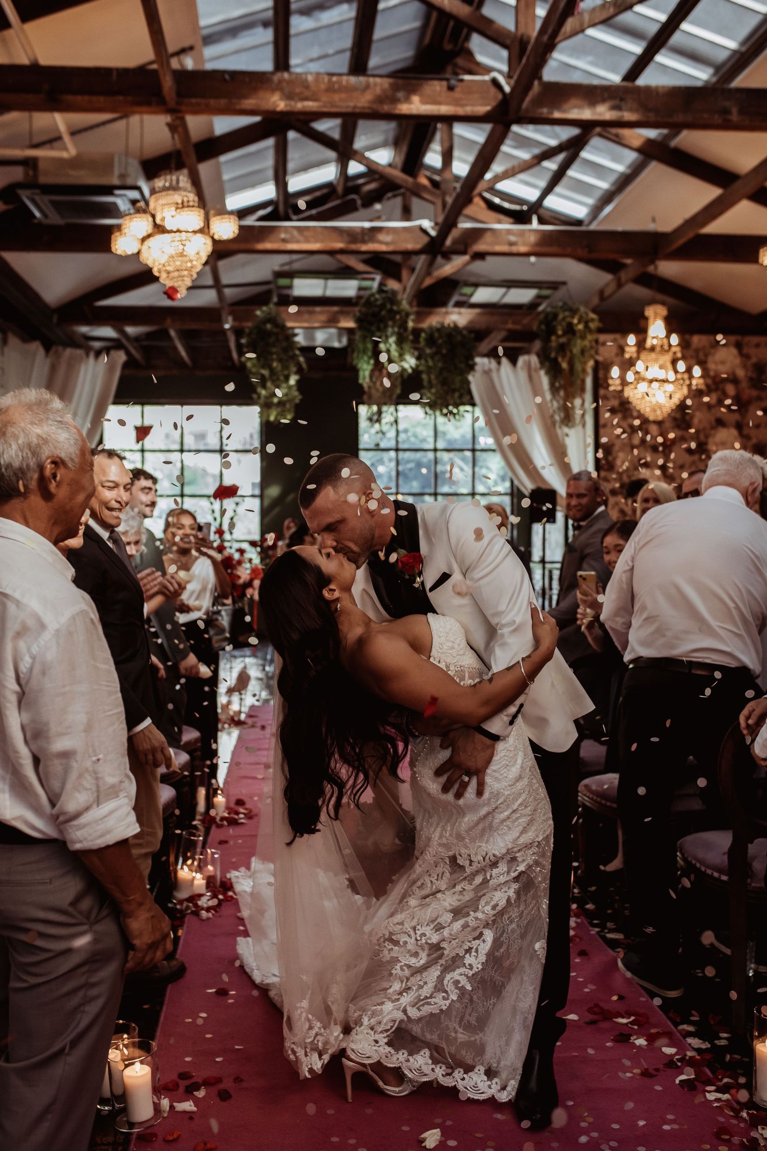 Couple dances at a rustic indoor wedding reception with chandeliers and exposed beams while guests celebrate around them.