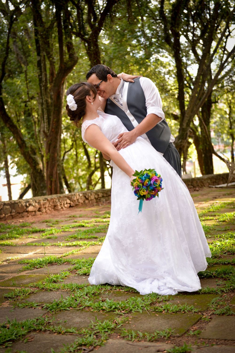 Bride and groom share a romantic kiss outdoors, surrounded by lush trees, as bride holds a colorful bouquet.