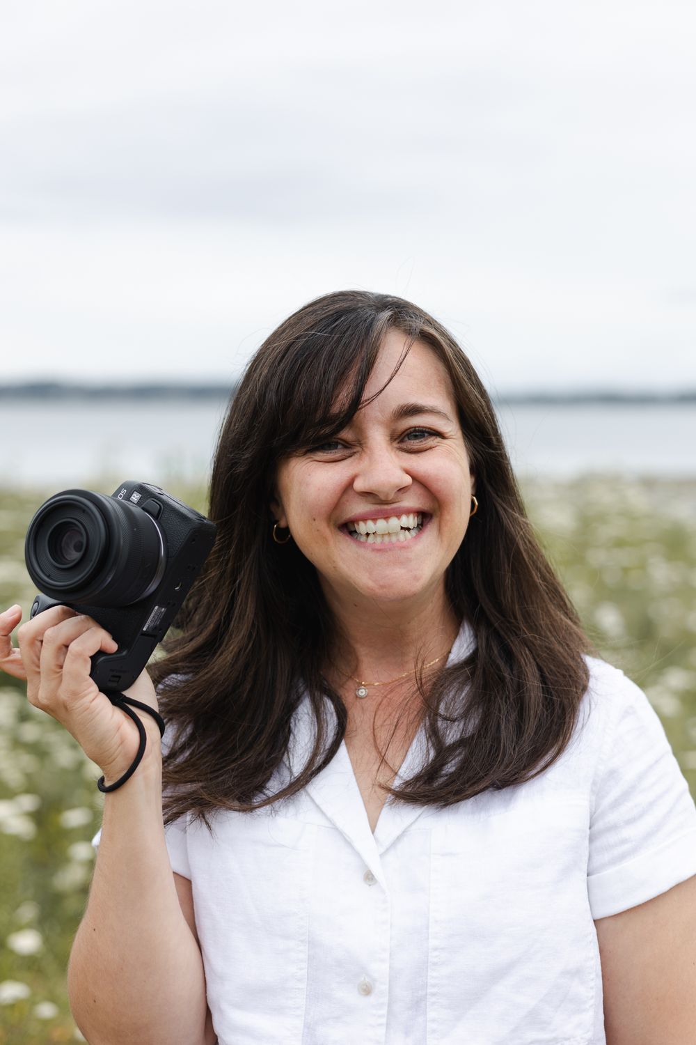 A photographer in a white shirt smiles while holding a camera against a bright outdoor background.