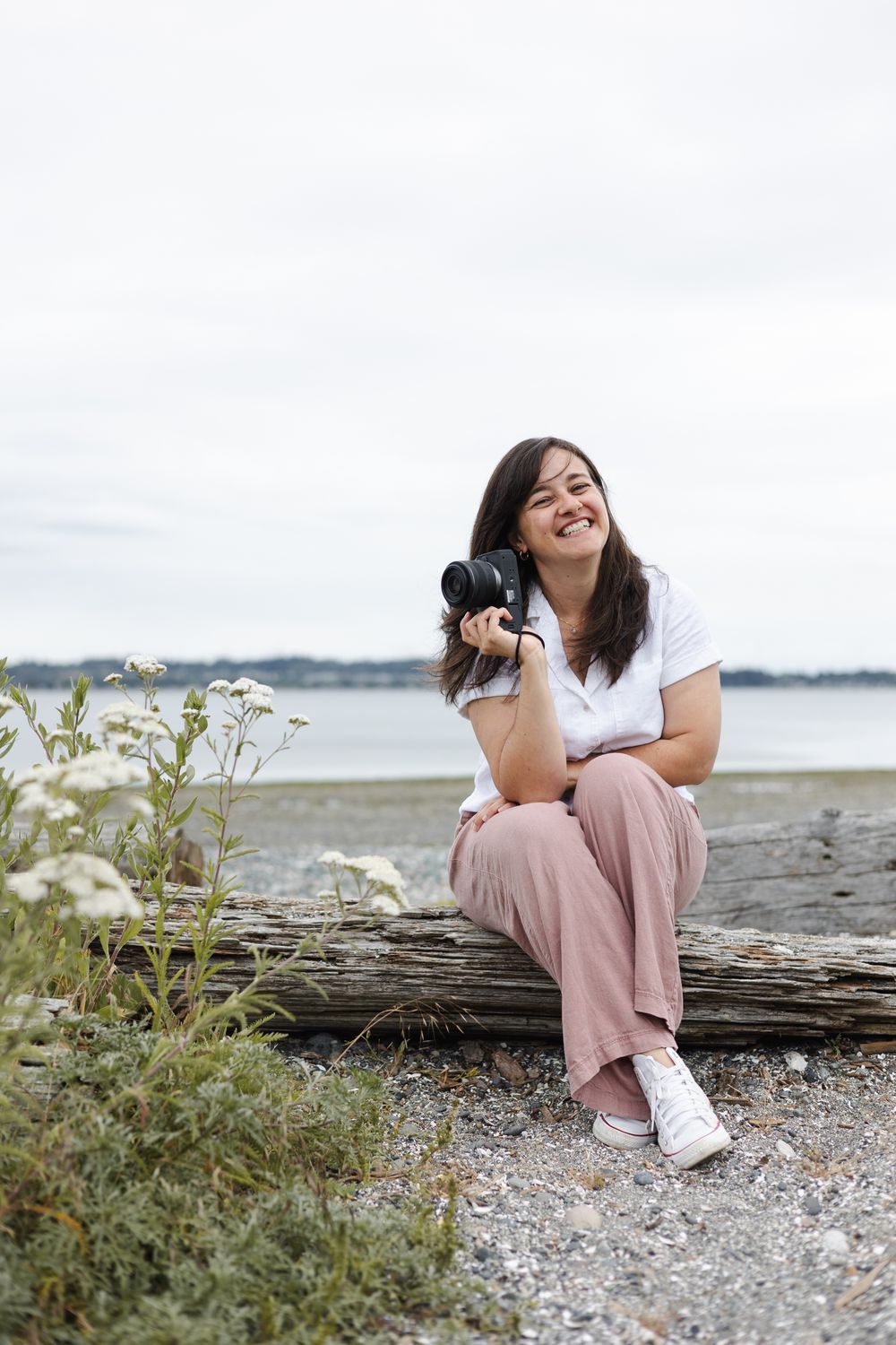 Person in pink pants and white shirt sits casually on driftwood at a peaceful beach with wildflowers nearby.