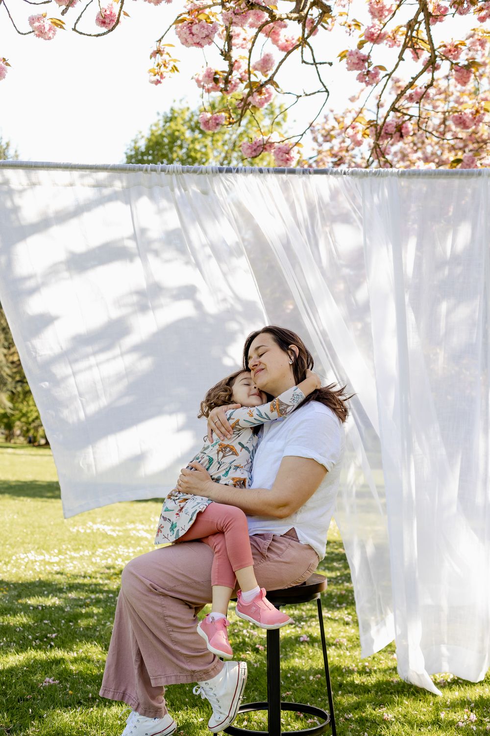 Mother and child share tender moments among white linens drying on clotheslines in a sunny spring garden.